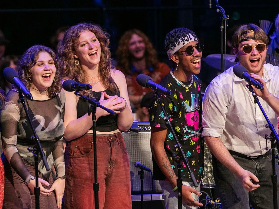 Four Skidmore College students sing together on stage, smiling and laughing under concert lights, during a live music performance with microphones and instruments visible in the background.