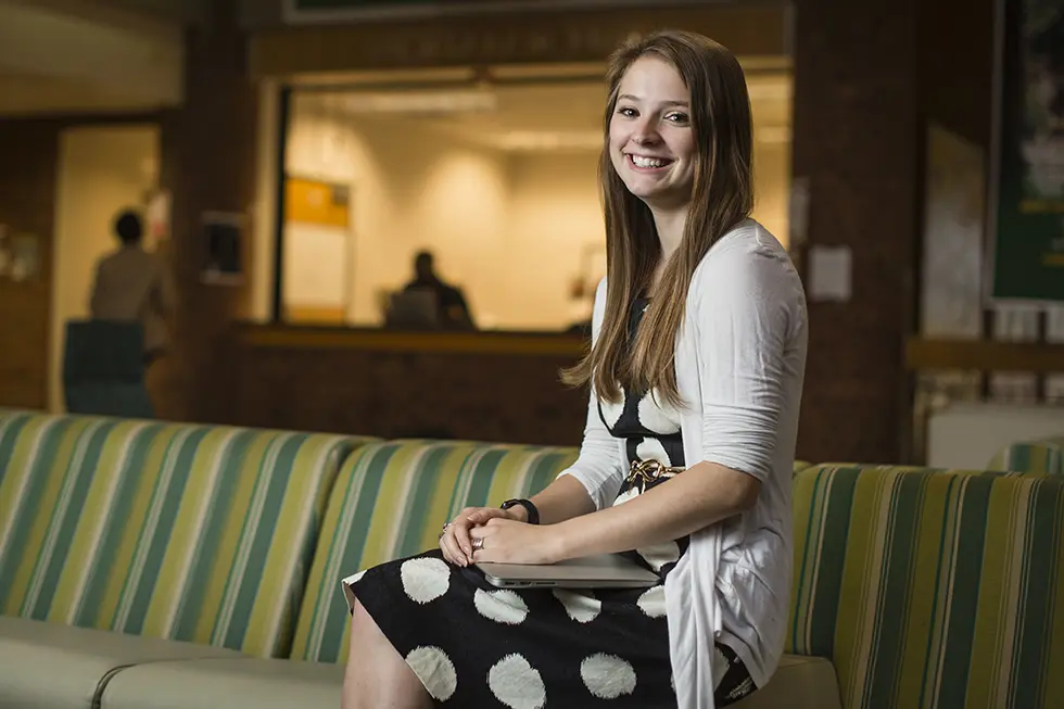 A student sits on a striped green and yellow couch in a campus lounge, smiling at the camera