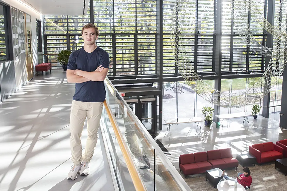 A student stands confidently with arms crossed in a modern, sunlit campus building with large windows and metal sculpture art hanging overhead.