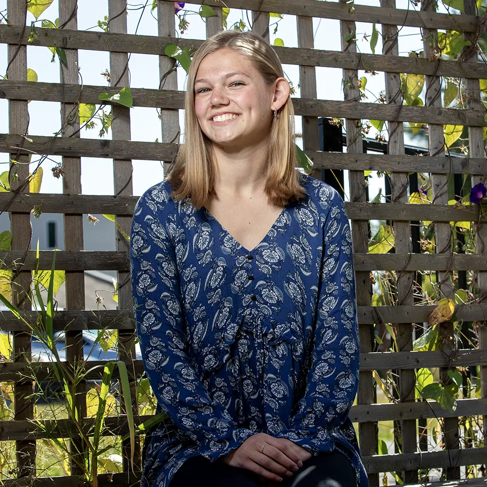 A student sits in front of a wooden trellis covered in green vines and flowers. She wears a blue patterned blouse and smiles in the natural light, with a relaxed and cheerful expression.
