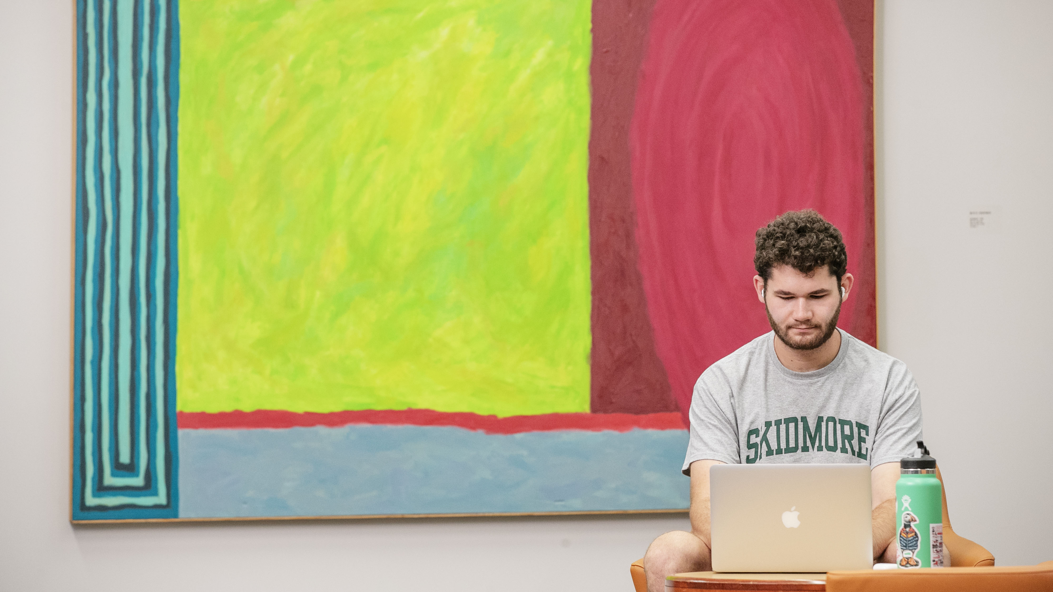 Milo Flamenbaum ’26 sits beside a large abstract painting in Lucy Scribner Library, working on a laptop.