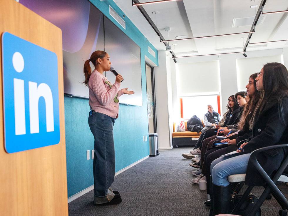 A presenter stands next to a podium with the LinkedIn in logo on the front, at LinkedIn headquarters with green wall and large screen