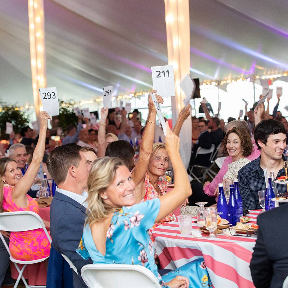 Event attendees sit at tables under a lighted tent raising numbers in the air for a game