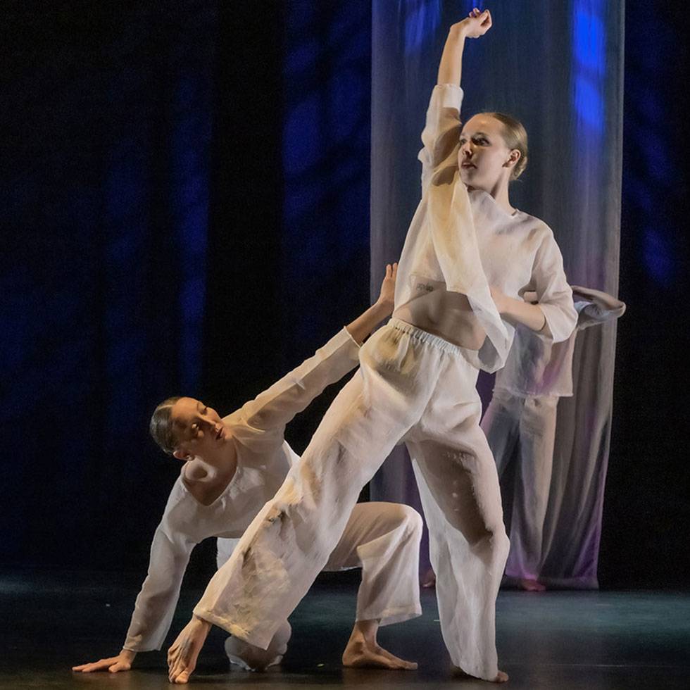A dancer in a white flowing shirt and pants stretches her arm in the air with a leg extended out, while another dancer below reaches up to her.