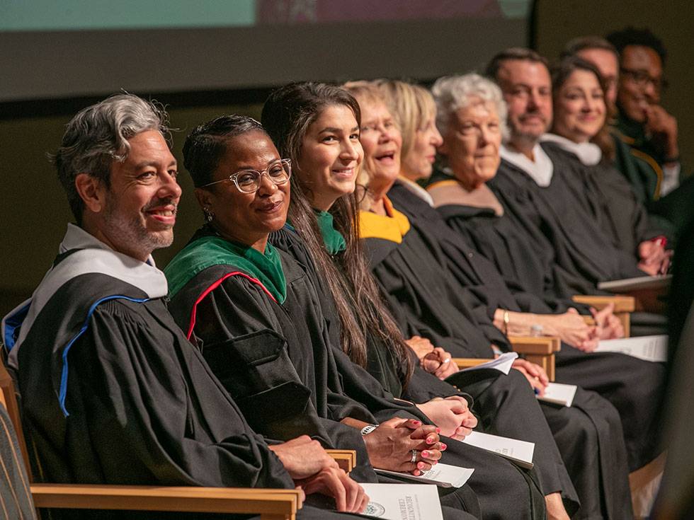A line of alumni award recipients sit on stage smiling while wearing regalia at the annual alumni awards ceremony in Zankel Music Center