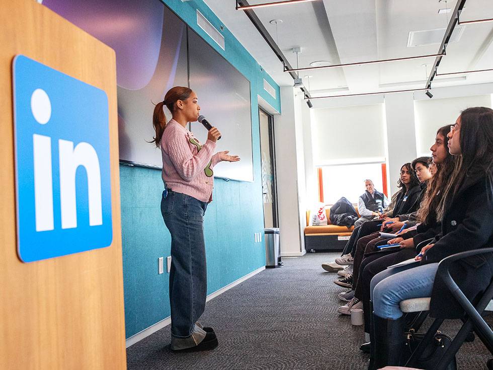 A women stands at the front of a seated group with a LinkedIn logo in blue to the right of her, while speaking to them in a microphone