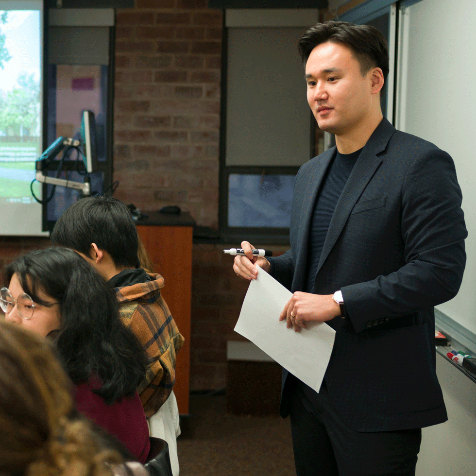 Professor Joowon Park lectures in a classroom while holding a piece of white paper in his hand, with students in the foreground