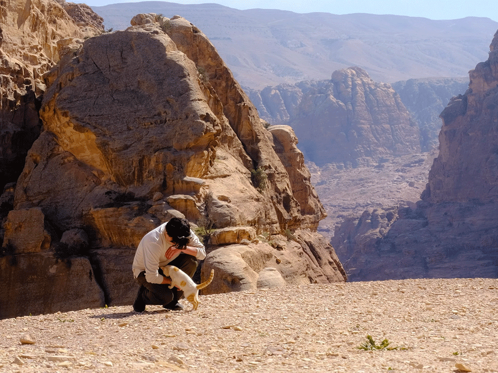 Student crouches down in a mountainous desert landscape to greet a white and orange cat