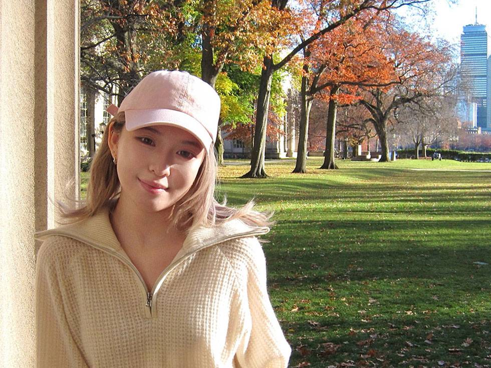 A student stands on campus in autumn, with colorful trees, a grassy lawn, and a city skyline in the background.