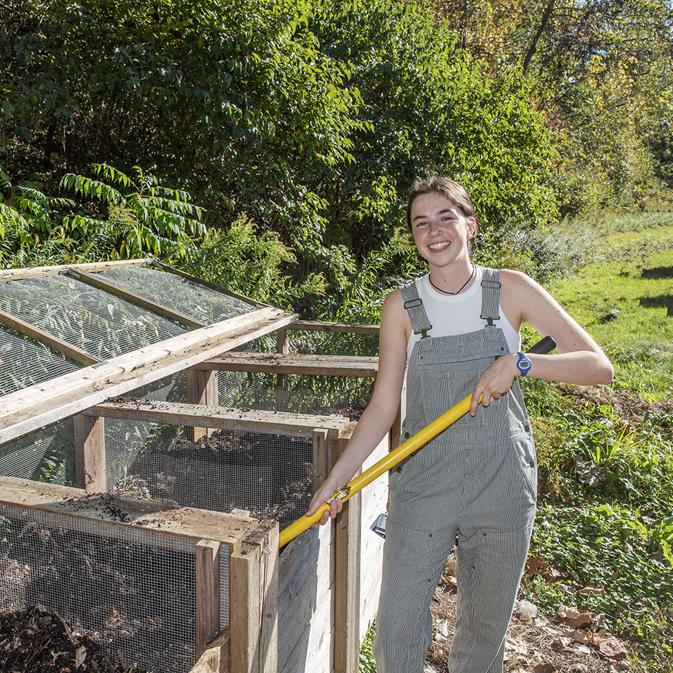 A person wearing light gray striped overalls and a white sleeveless shirt smiles while using a yellow-handled rake at an outdoor composting area. Wooden compost bins with mesh panels and glass covers are visible beside them, surrounded by green trees and grass under bright sunlight.