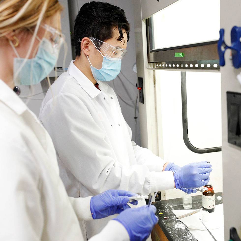 Two chemistry students work in a lab, wearing white lab coats, blue gloves, masks, and safety goggles.