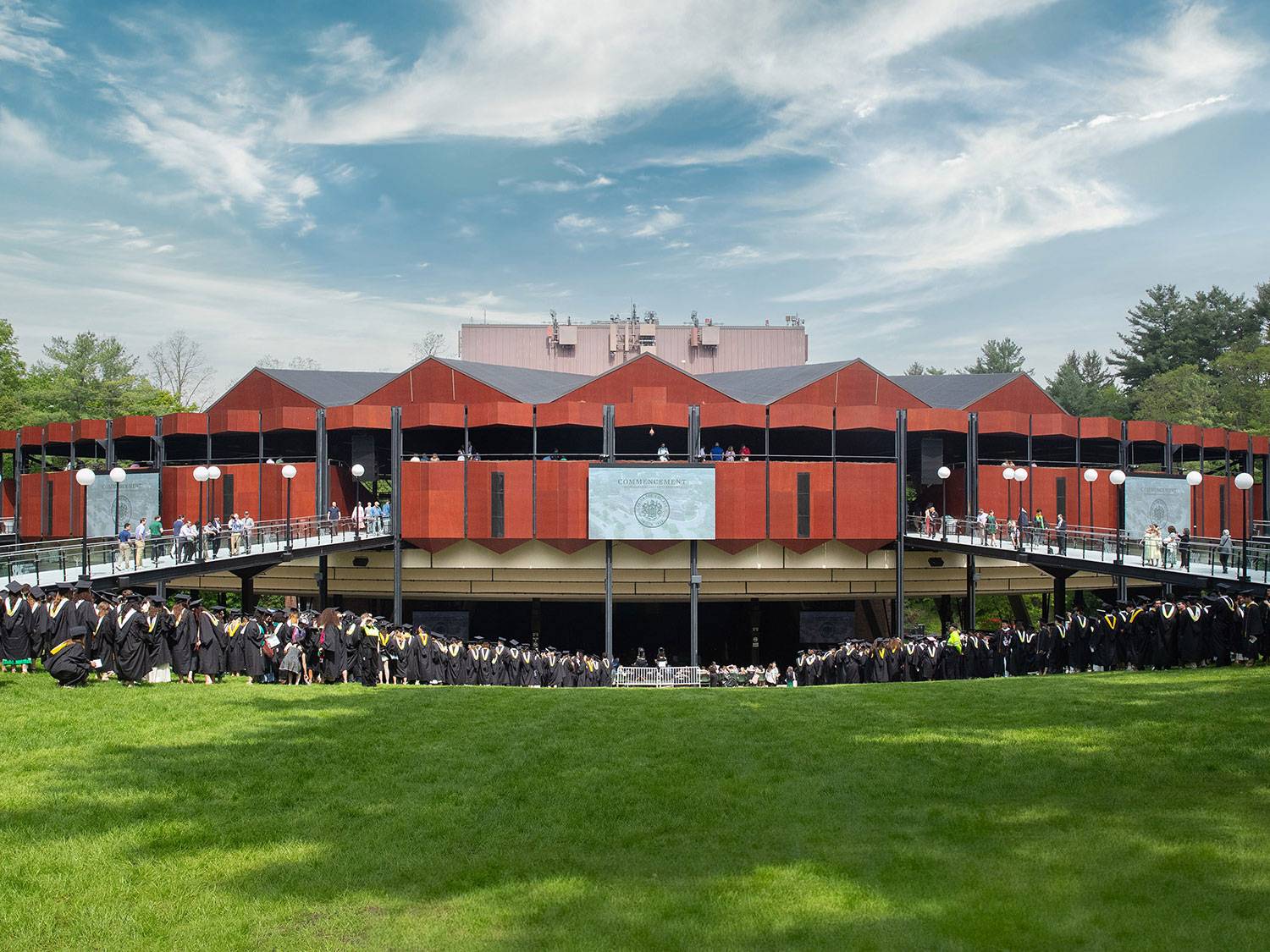 A beautiful blue sky day, showing SPAC's ampitheater with a Skidmore Commencement screen centered and students in commencement regalia  lined up on either side