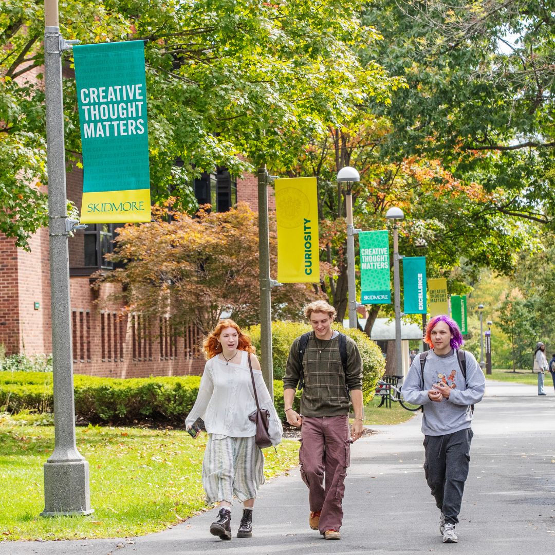 Three students walking along a tree-lined campus path beneath green and yellow Skidmore banners reading “Creative Thought Matters” and “Curiosity.”