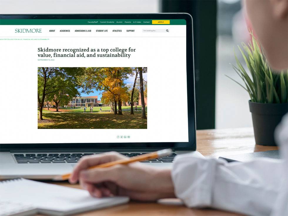 Image of a laptop computer with a Skidmore news story pulled up, and including a person in front of the screen taking notes with a pencil.