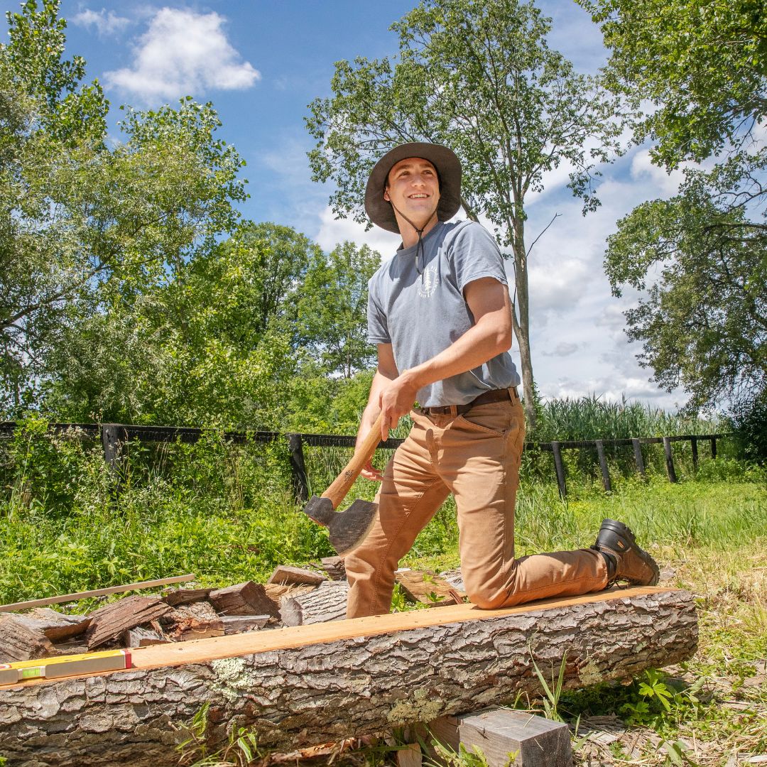 Student wearing a wide-brim hat kneeling outdoors and splitting a log with an axe in a grassy, wooded area under a blue sky.