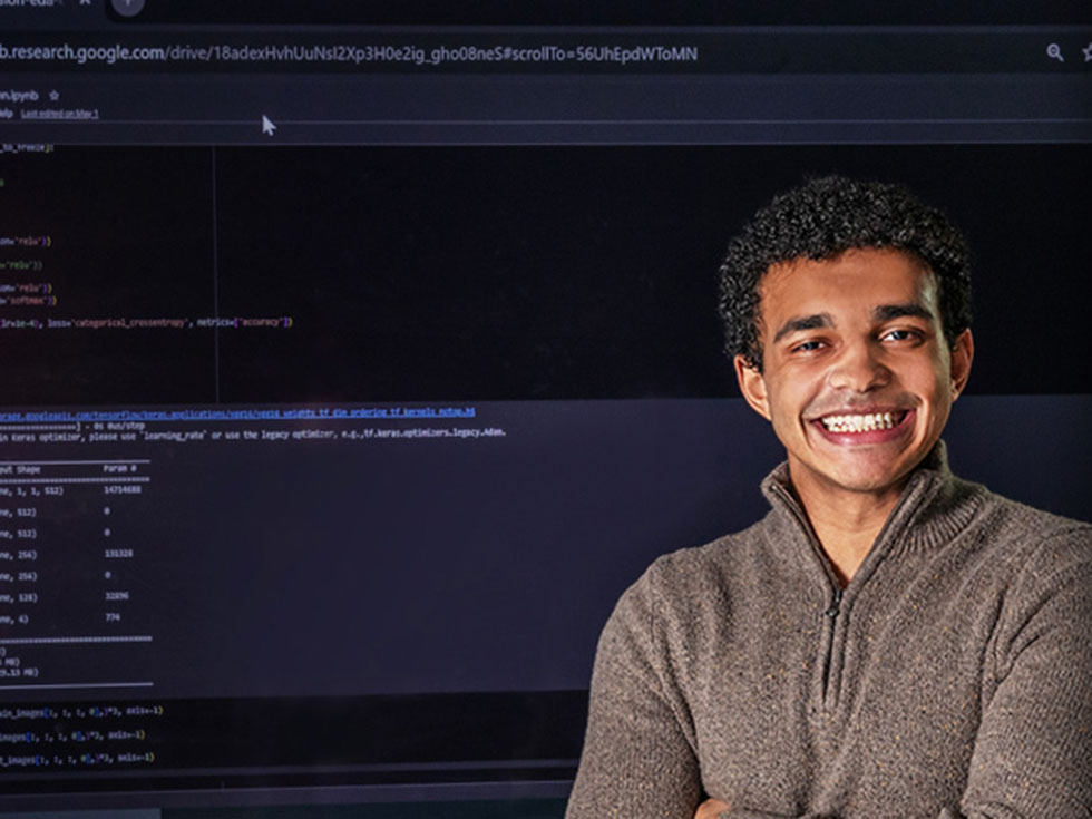 A smiling young man stands confidently in front of a large screen displaying computer code and a Google Colab interface. He wears a brown quarter-zip sweater and poses with arms crossed, conveying enthusiasm for computer science and programming.