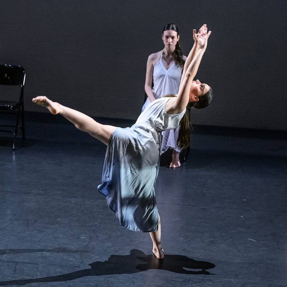 Dancer performs on a dark stage with arms thrown in the air while she looking up and extends one leg out into a point