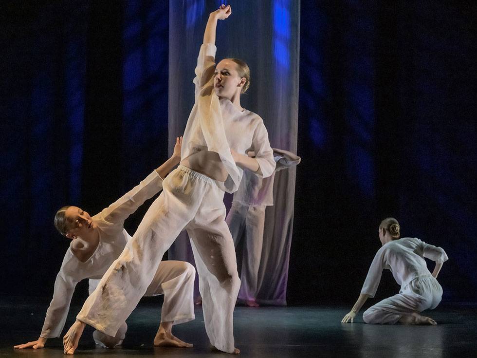 Dancer performs on stage in white flowy outfit while reaching towards the sky with two other dances in dark background