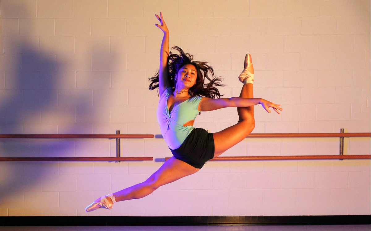 A dancer leaps gracefully in midair, wearing pointe shoes, a turquoise leotard, and black shorts, against a studio wall with ballet barres.
