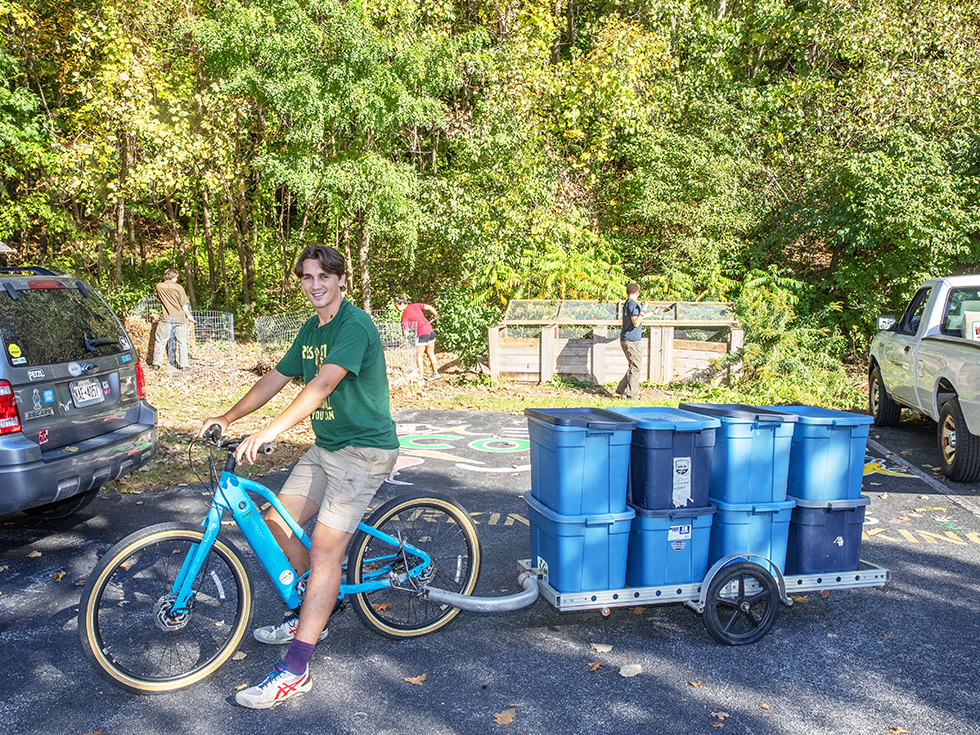 Zak Maxey rides a bike with a cart behind hauling recycling boxes.