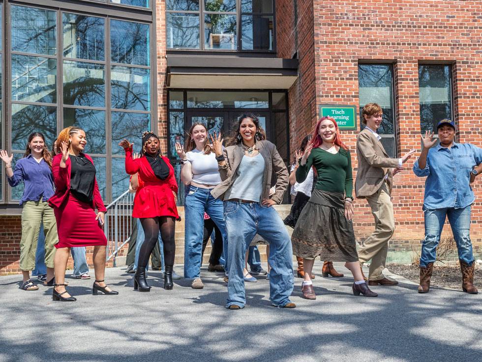A group of students dance outside a brick academic building with their right arm thrown into the air and smiling.