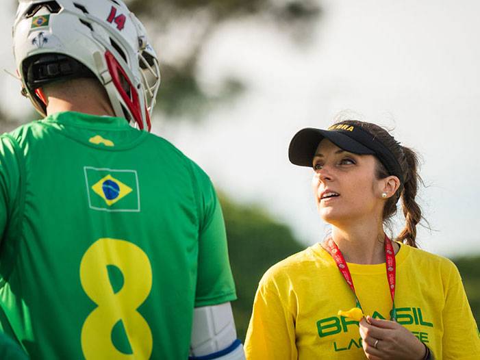 Cristina Velez talks with a Brazilian lacrosse player. She is wearing a yellow team shirt and black hat, and the player is in their green uniform with a white helmet.