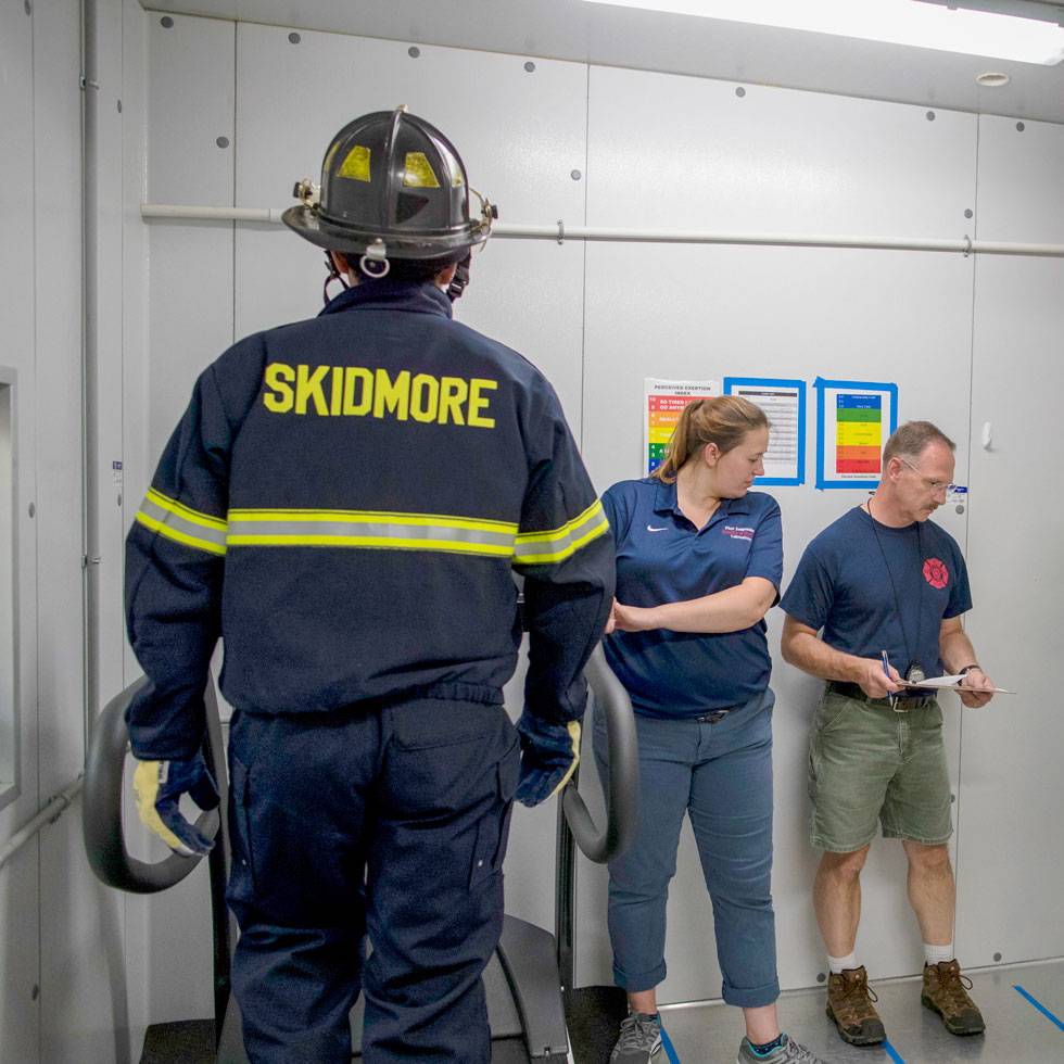 A firefighter stands in their uniform that is black with neon yellow text that says "Skidmore," and two other people stand against the lab wall taking research notes.. 