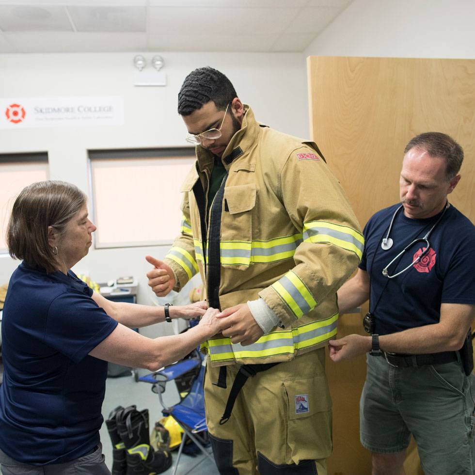 Three individuals stand in a lab, preparing a firefighter for a research study.