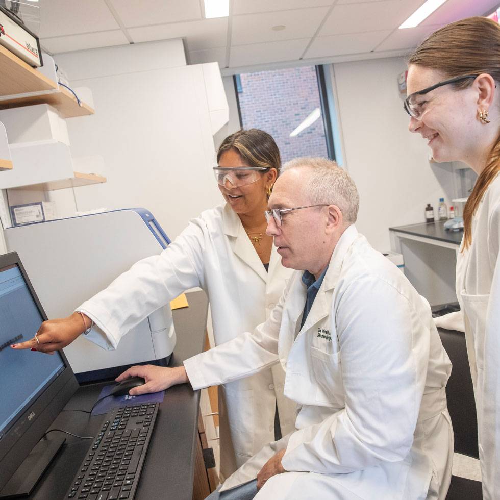 Two students and a professor wearing white lab coats look at a computer screen in a lab.