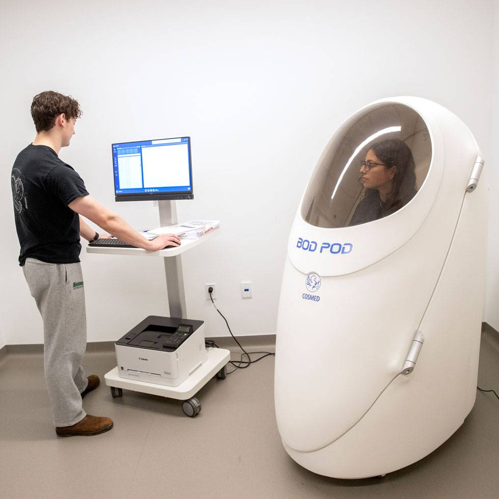 Two students work in a lab that includes one working on a computer, and the other in a Bod Pod capsule.