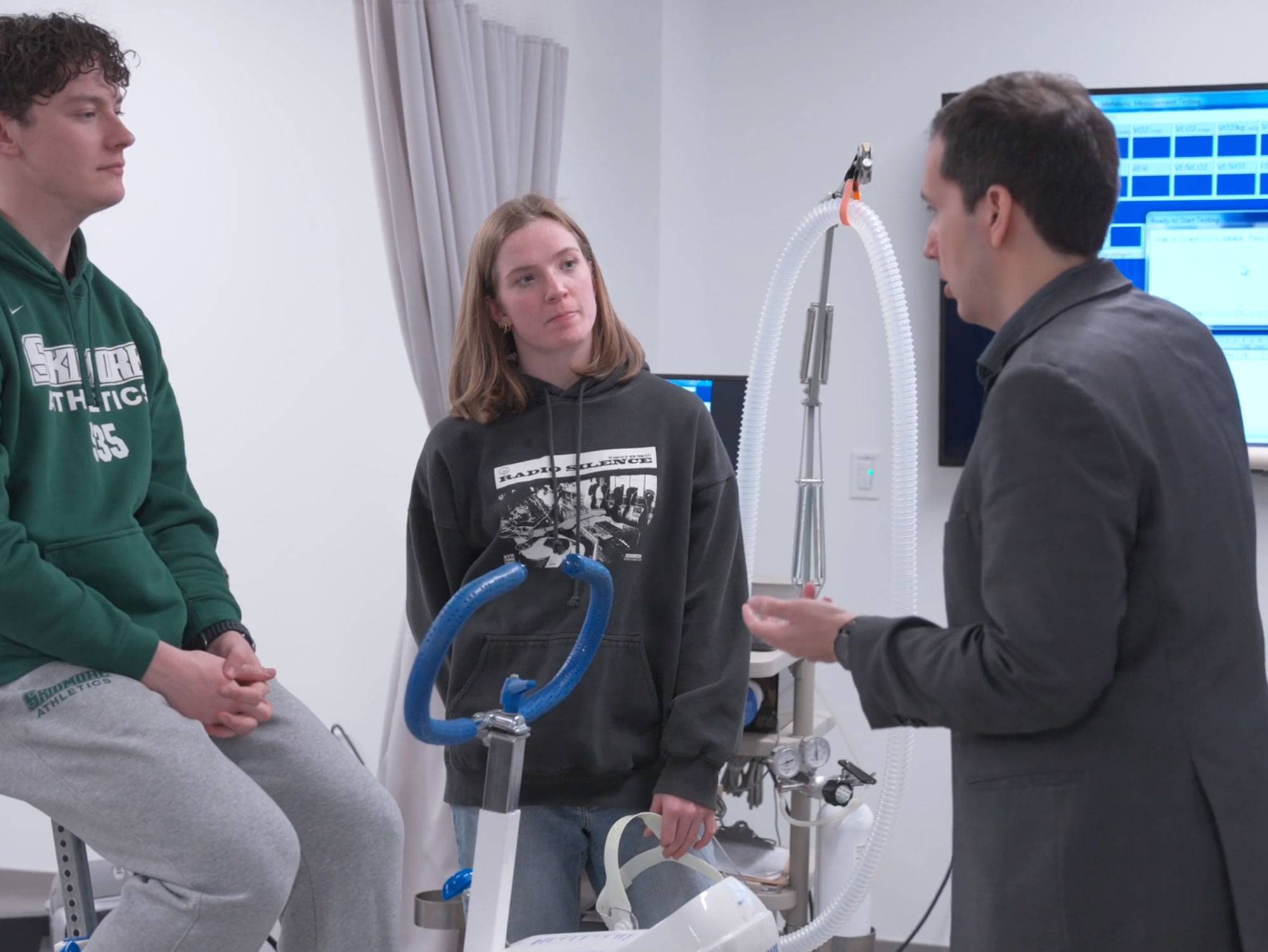 Three people stand talking in a white exercise lab. One is a on stationary bike, one is standing, and both individuals are listening to the third person who has their hands out speaking.