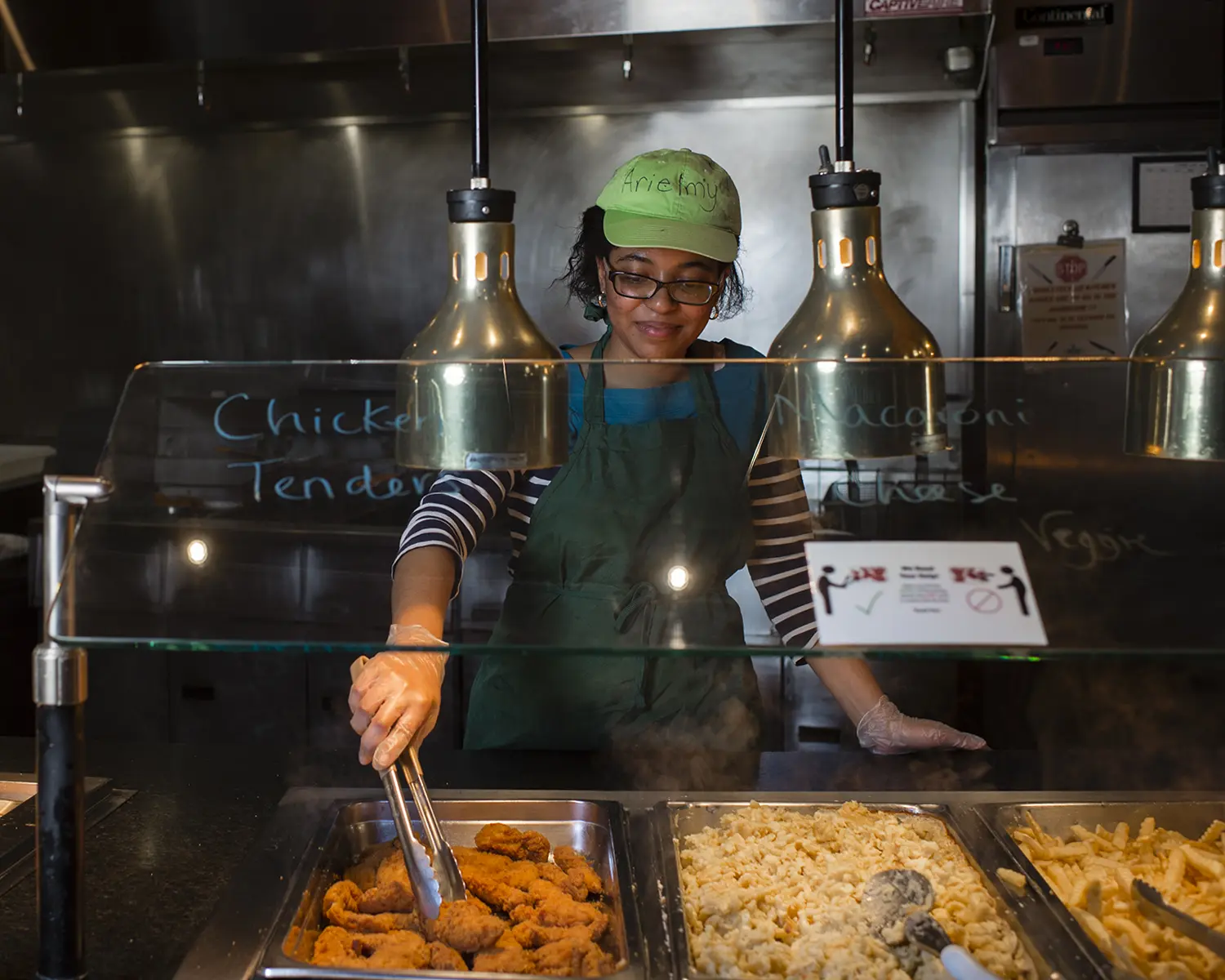A student working in the dining hall serves chicken fingers and macaroni and cheese.