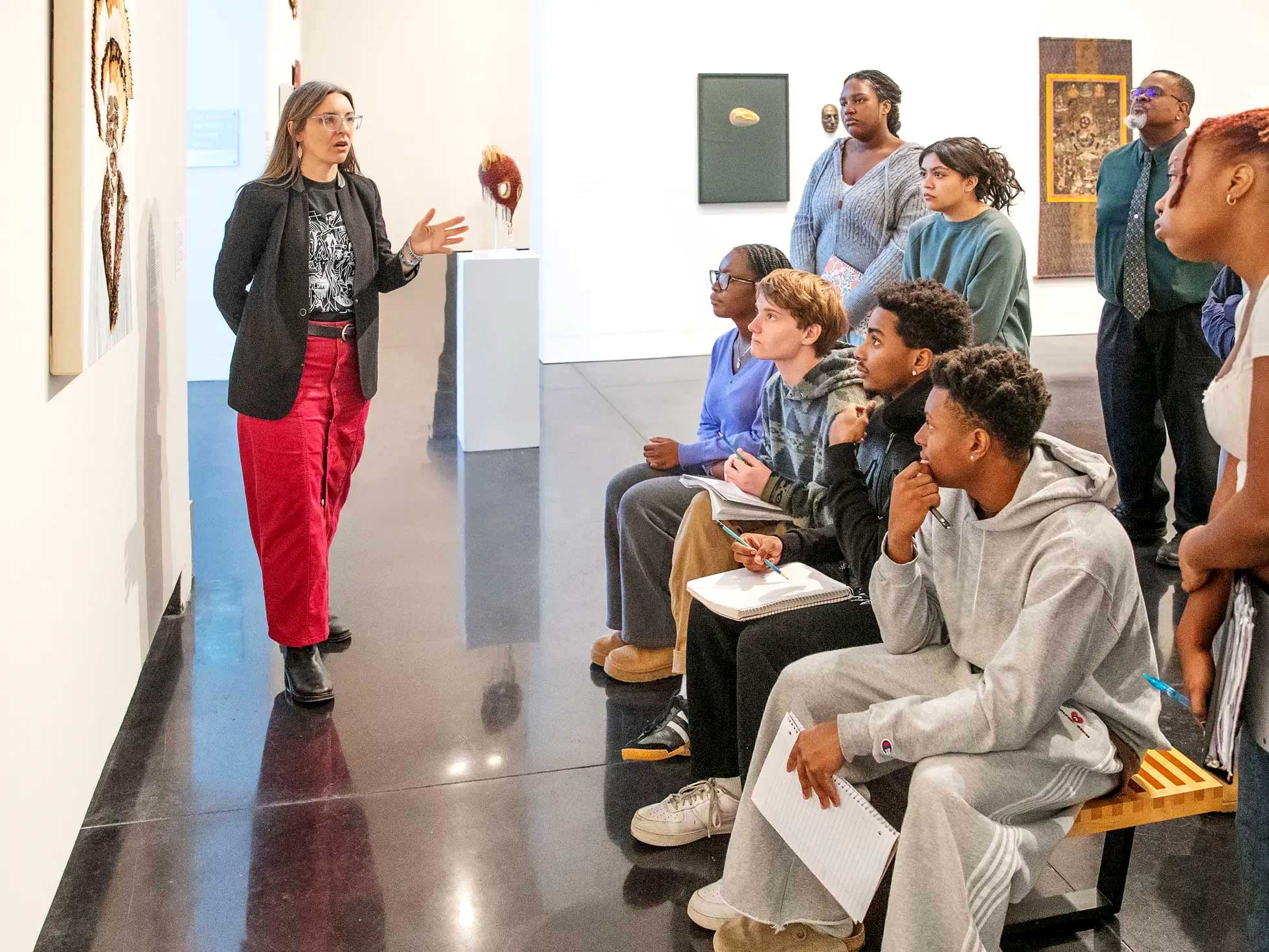 A faculty member leads a gallery discussion with students taking notes while viewing artwork in a museum classroom.