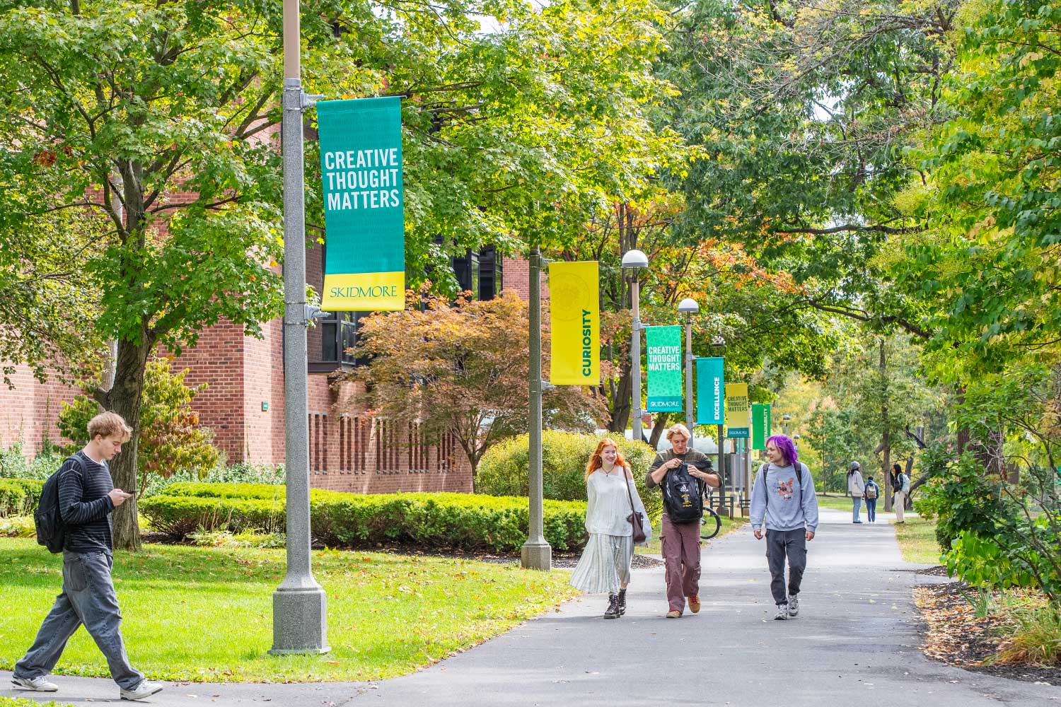 tudents walking along a tree-lined campus path with Skidmore banners reading “Creative Thought Matters” and “Curiosity” hanging from lampposts.