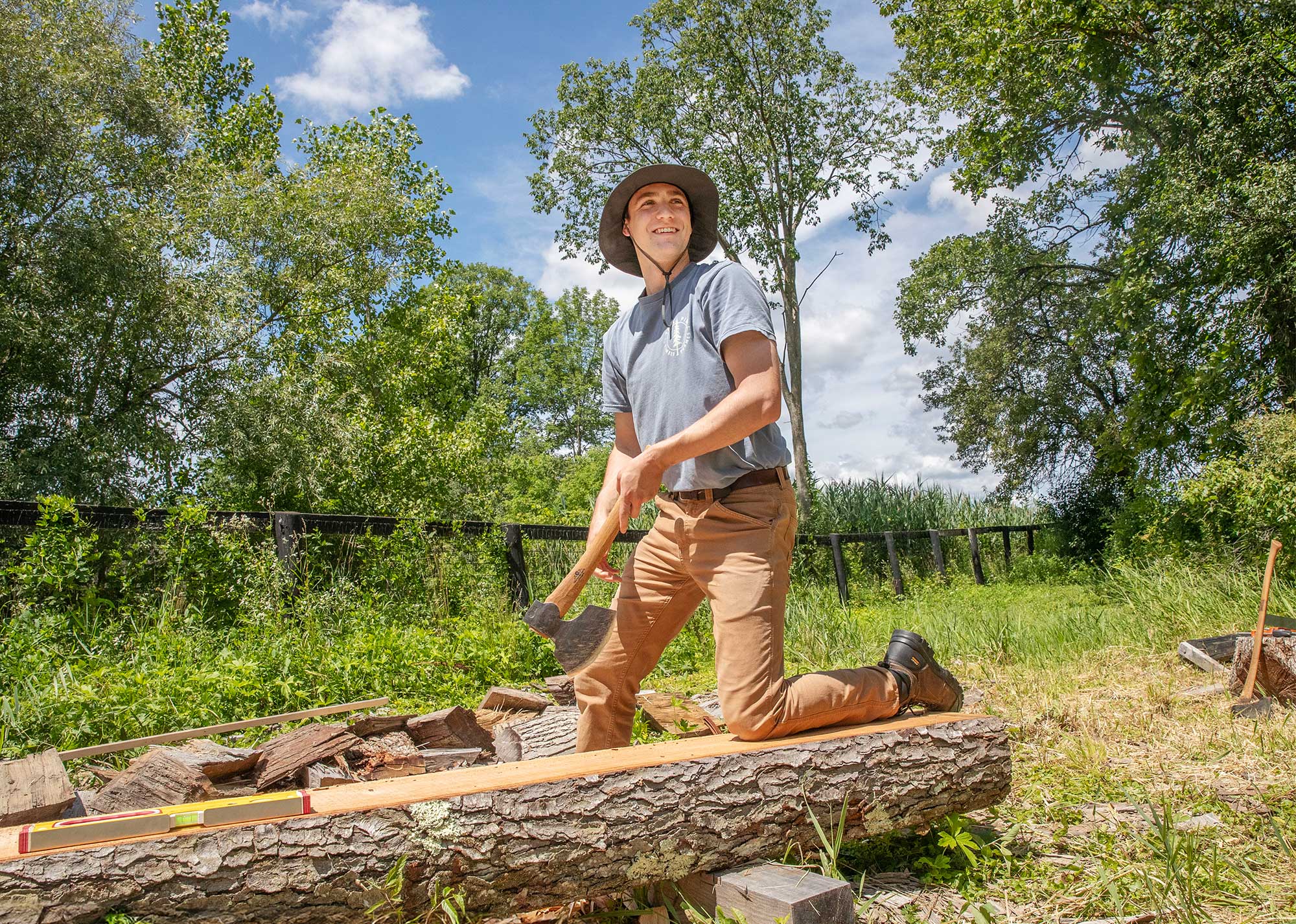 Student wearing a wide-brim hat kneeling on a log and splitting wood with an axe in a grassy, wooded area under a bright blue sky.