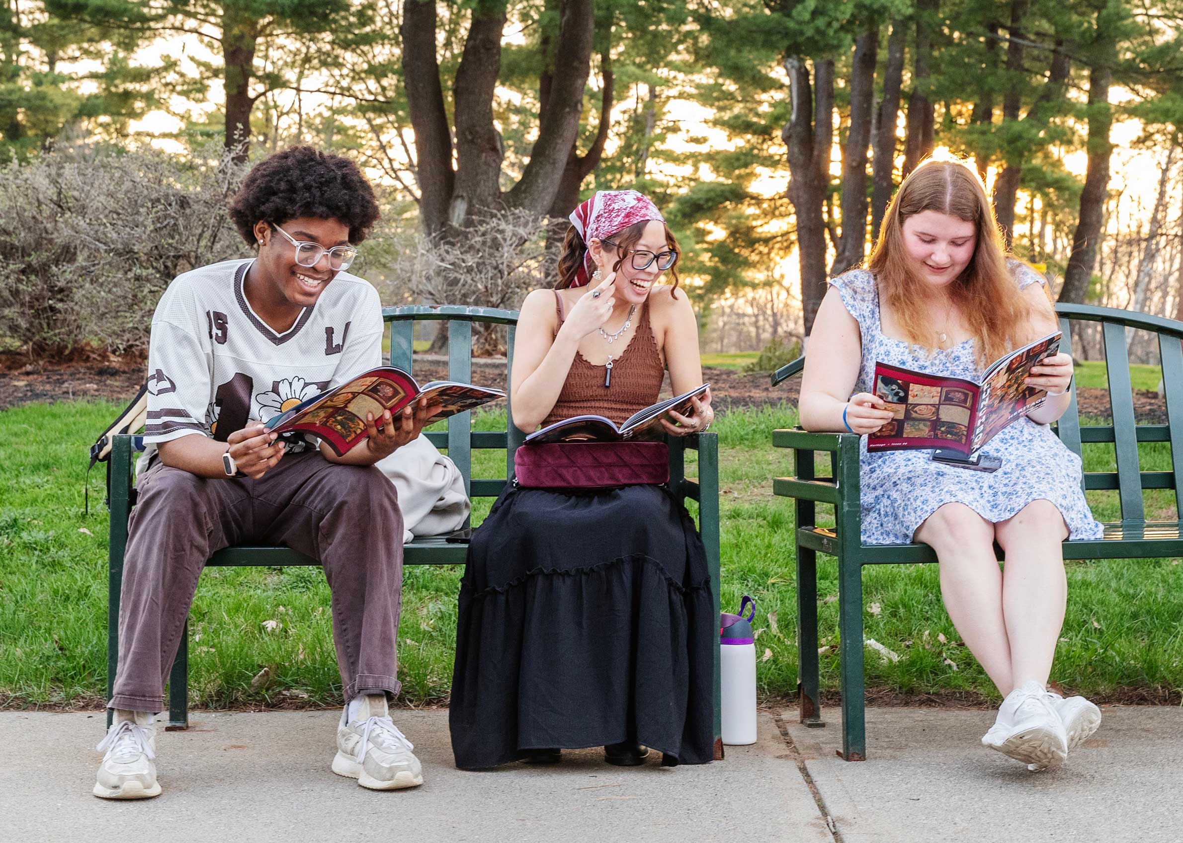 Three students sit on benches outdoors, smiling and reading magazines together at sunset.