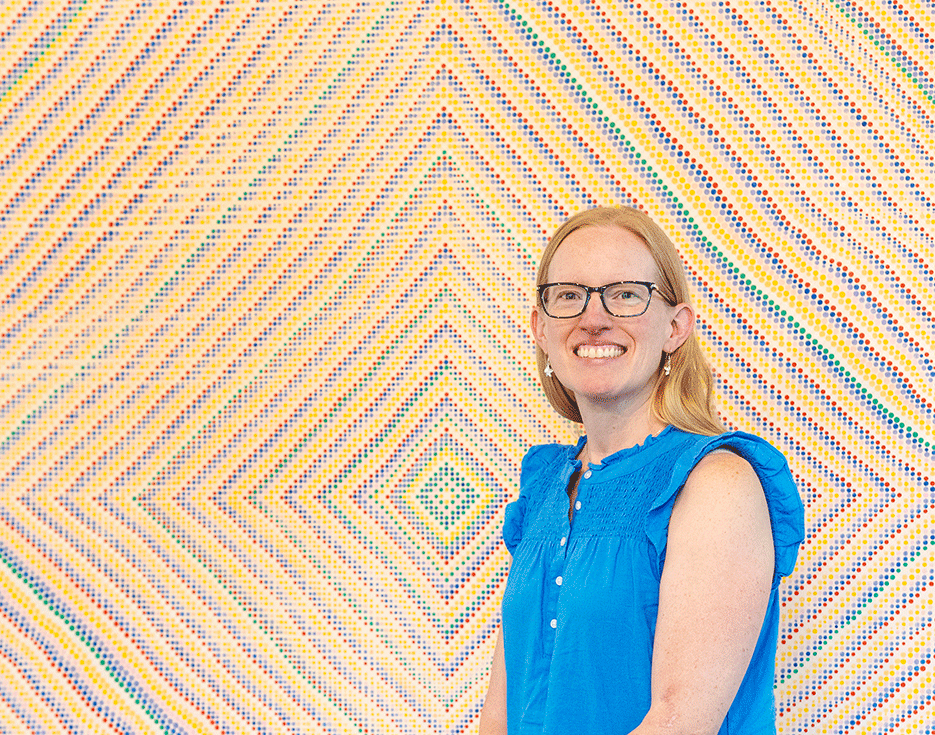 Professor Rachel Roe-Dale sits smiling wearing a bright blue shirt with a colorful geometric painting in the background