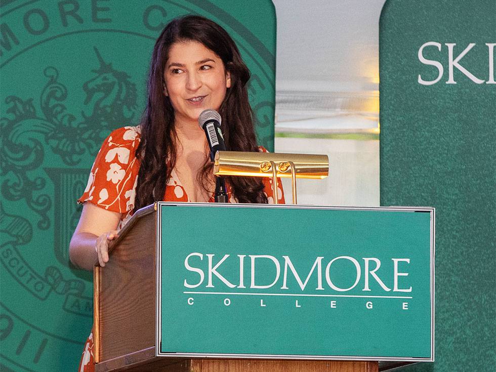 Alina Bazarian stands on stage at a Skidmore podium speaking into a mircophone and wearing a red dress with white floral print.