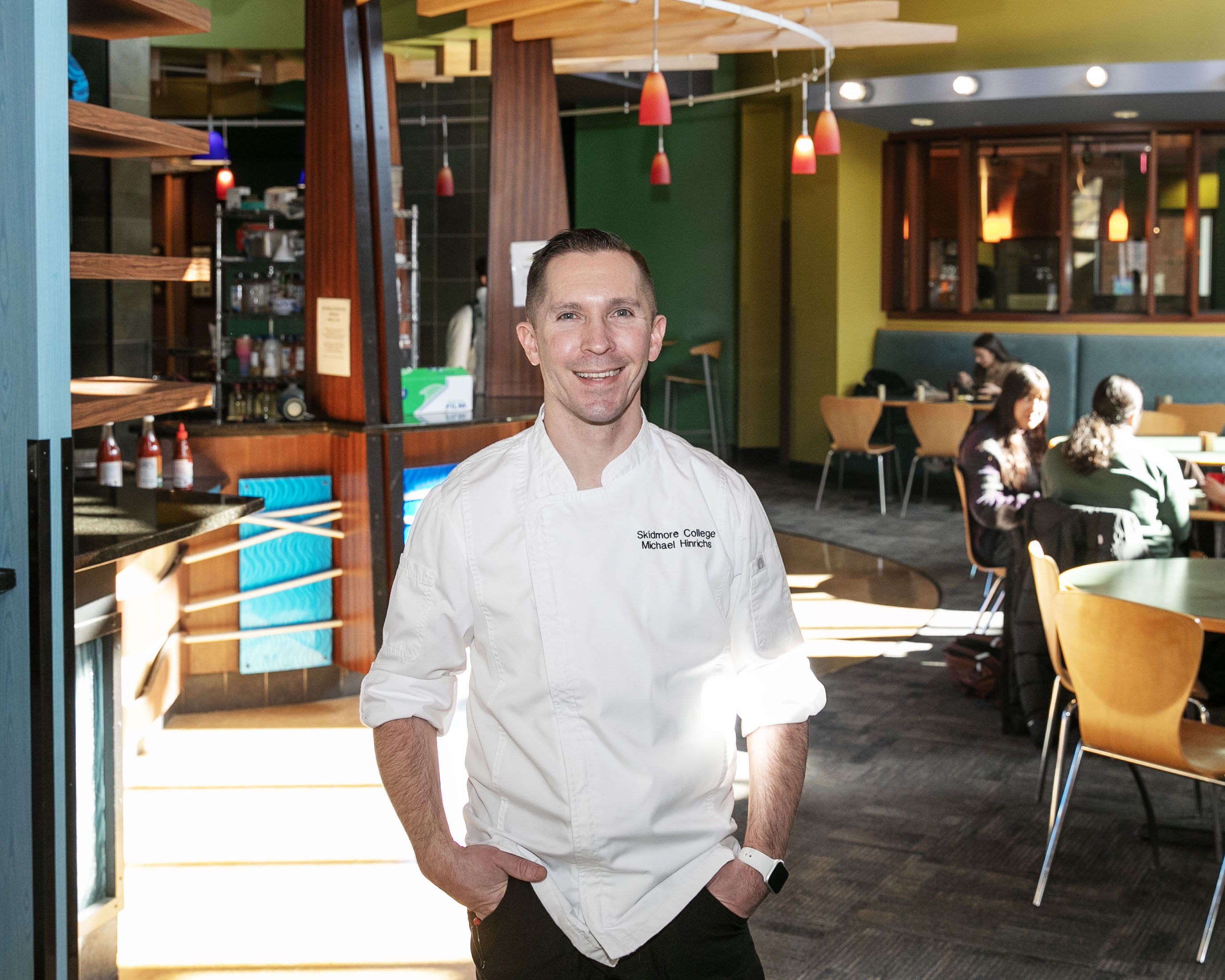 Michael Hinrichs stands, smiling in Murray-Aikins Dining Hall. A few students sit at tables in the background.