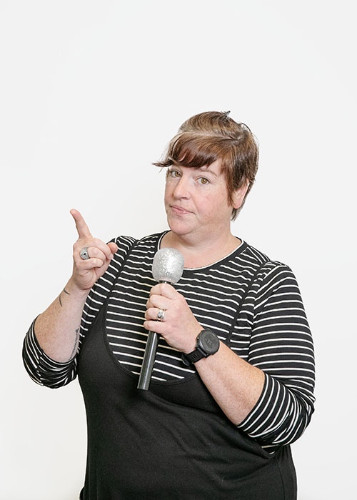Beck Krefting Beck Krefting, wearing a black and white striped long sleeve shirt under a black dress, holds up an admonishing finger and a sparkly microphone