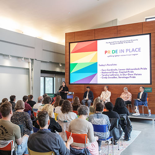 A+crowd+sits+on+multicolored+chairs+in+the+Wyckoff+center%2C+watching+a+stage+with+five+people+and+a+large+screen+with+%22Pride+in+Place%22