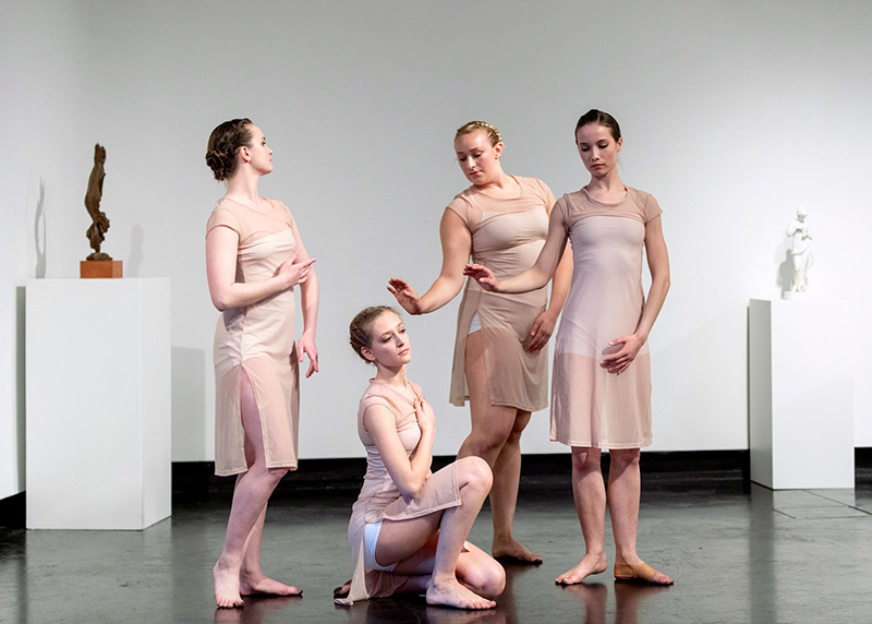 Four dancers pose in tan-colored dresses among several sculptures in the Tang Teaching Museum.