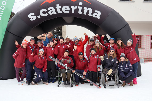 A large group of U.S. Paralympic Alpine Ski Team athletes and staff in red jackets cheer and raise their arms.