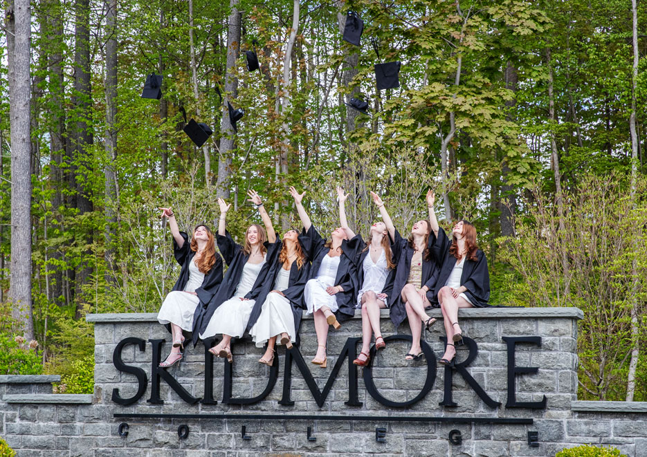 A group of graduates in caps and gowns celebrate by tossing their graduation caps into the air while sitting on a stone sign in front of a wooded backdrop at Skidmore College.