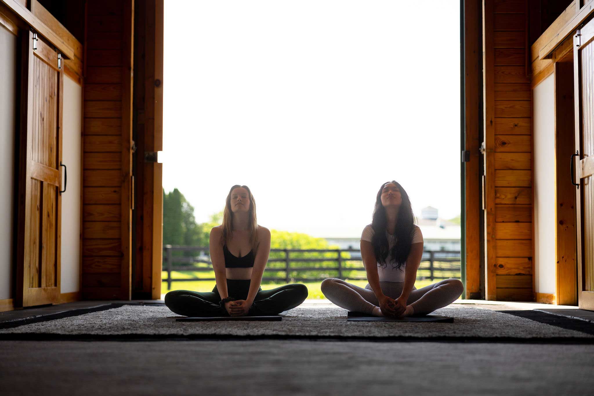 Two people practice seated yoga inside a bright wooden barn with open doors looking out to a sunlit field.
