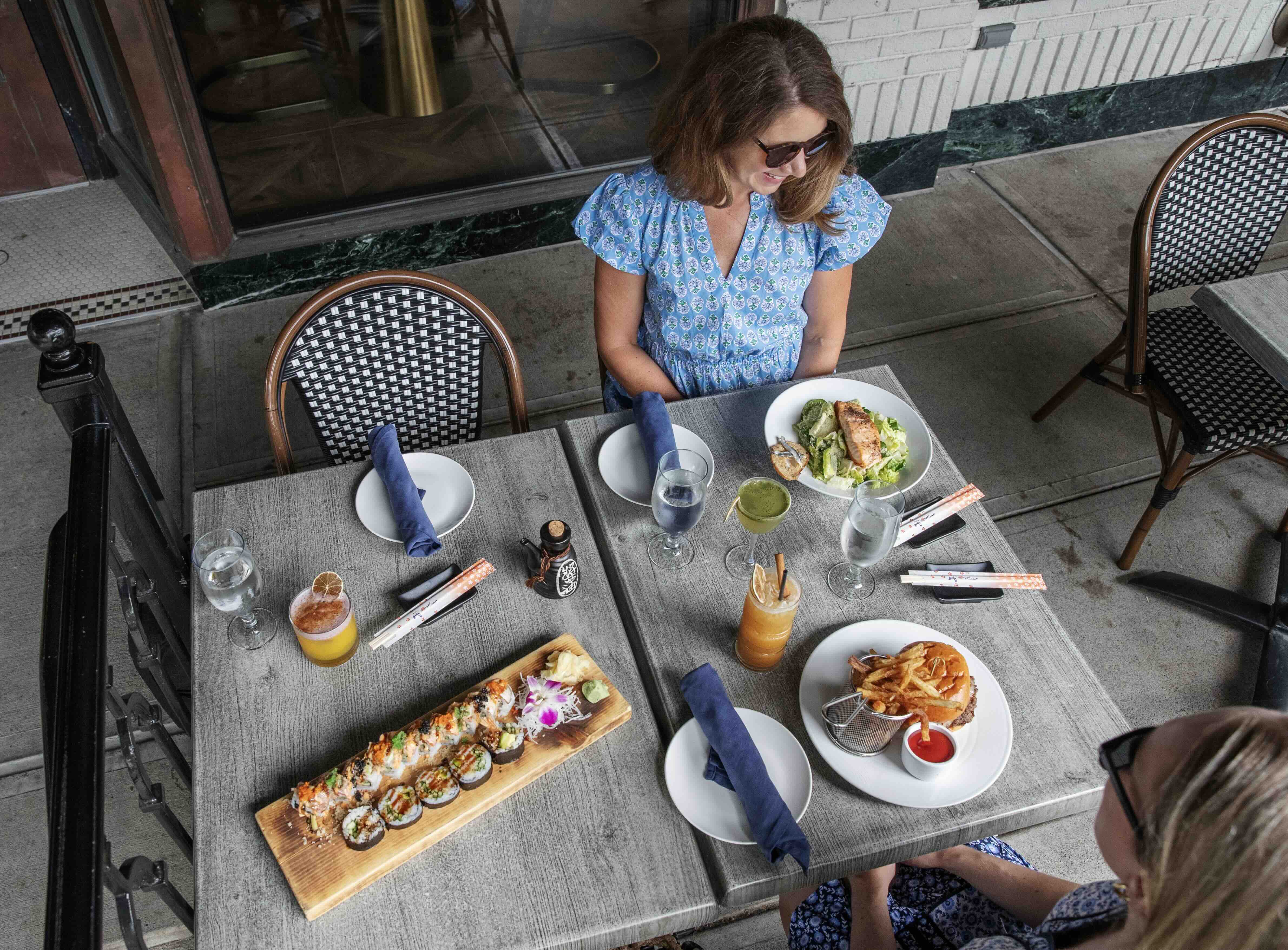Two women enjoy lunch at an outdoor restaurant table with sushi rolls, a burger and fries, salad, and colorful drinks.