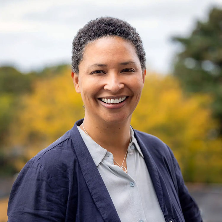 Person looks toward the camera and smiles, they are standing in front yellow and green fall foliage in the background.