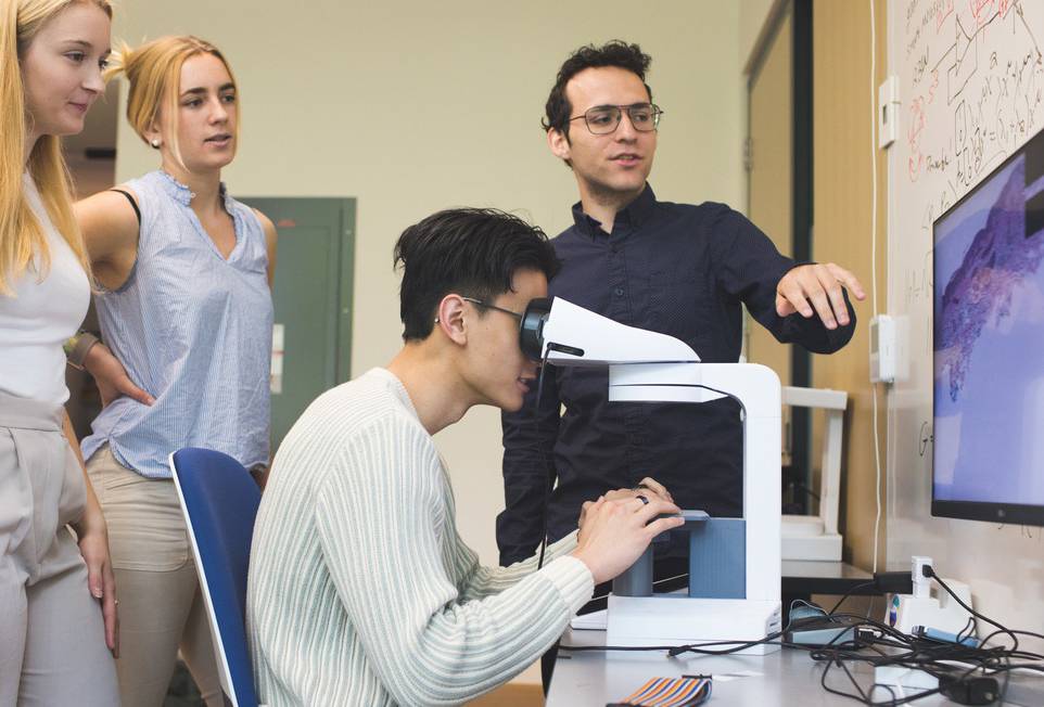 Alex Carney, Skidmore Class of 2022, explains the SmartScope, which could speed up clinical diagnoses by 50%, as a student looks through it and two others look on.