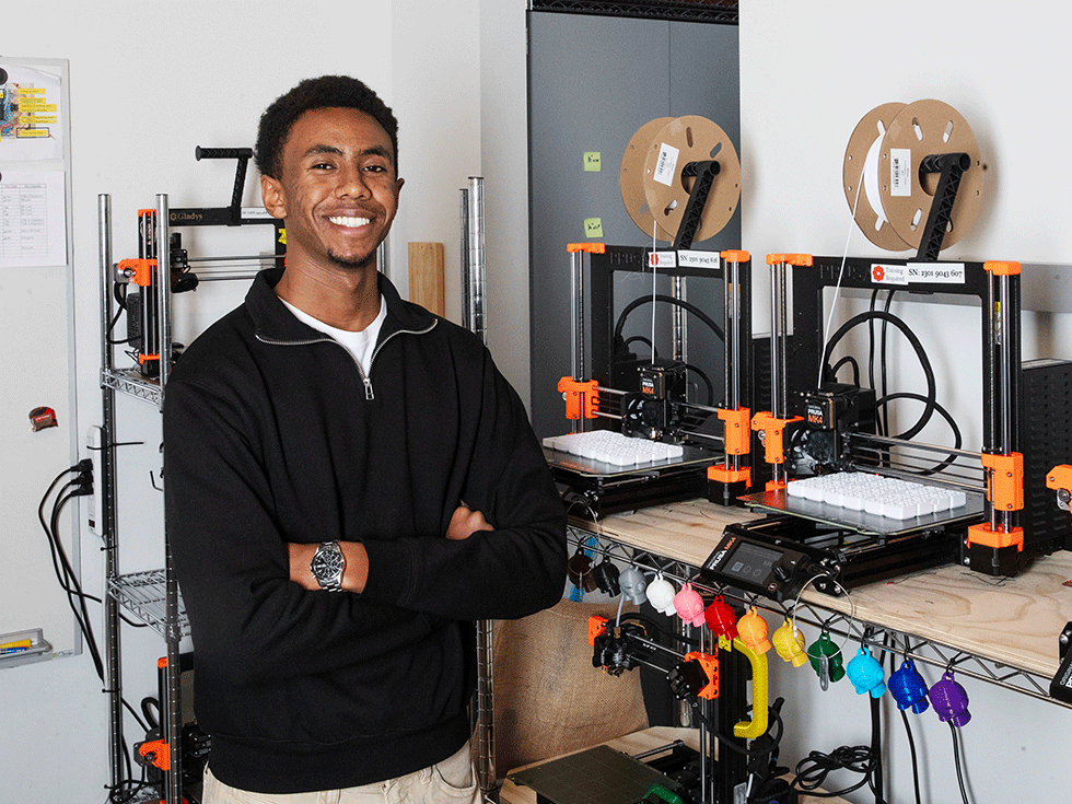 Isiah Karitanyi smiles with his arms folded in front of a 3-D printer in Billie Tisch Center for Integrated Sciences