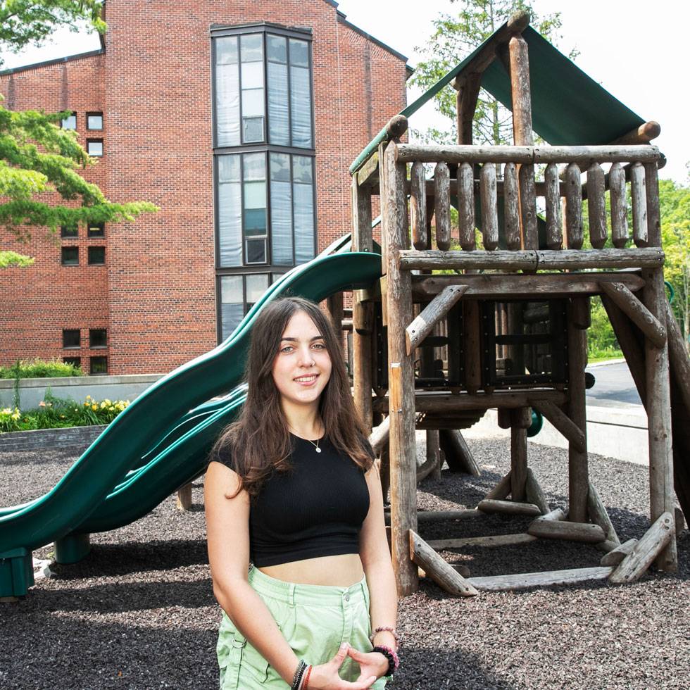Elena Tittel stands outside of a brick academic building with a wooden play structure in the background.